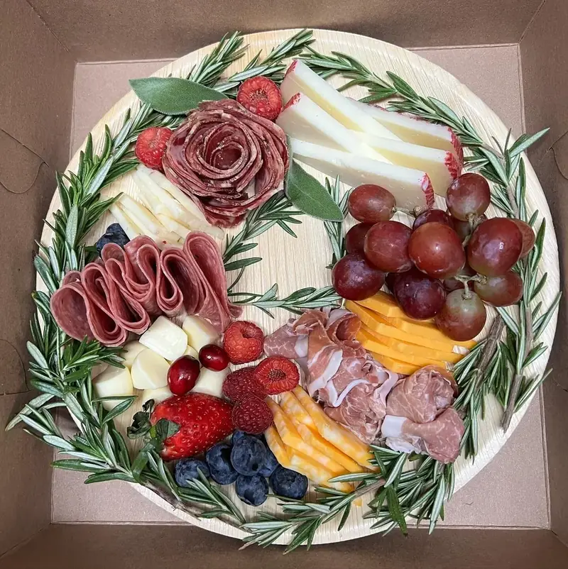 Round charcuterie wreath board with rosemary, salami roses, cheese, and fresh fruit from Lu's Cafe & Bakery